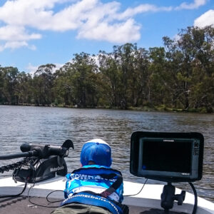 Aboard the boat, a figure sporting a Lowrance visor and a cool blue shirt takes in the serene vibe of the tree-lined river. The scene gets an upgrade with some serious kit on board, including the trusty Lowrance HDS unit—because why settle for just peaceful when you can have perfectly dialed-in peace?.
