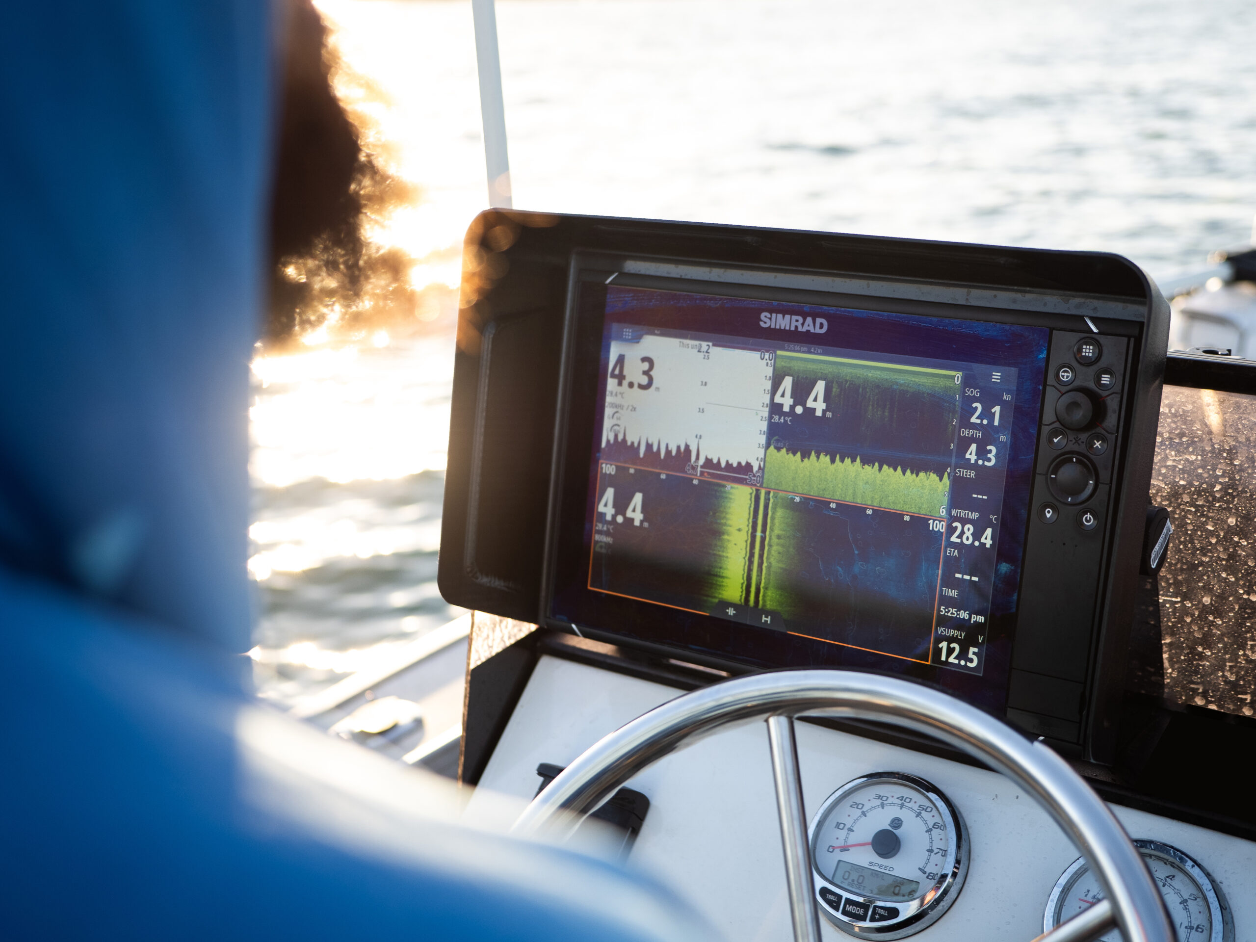 Rockin’ that blue hoodie, our boater takes the helm with style and precision. They’re deftly maneuvering their ride, all while tweaking their trusty Simrad Visor for a crystal-clear view of that sweet sonar data on the Simrad NSS EVO screen. Meanwhile, the water’s throwing its own shimmer party in the backdrop—pure boating bliss!.