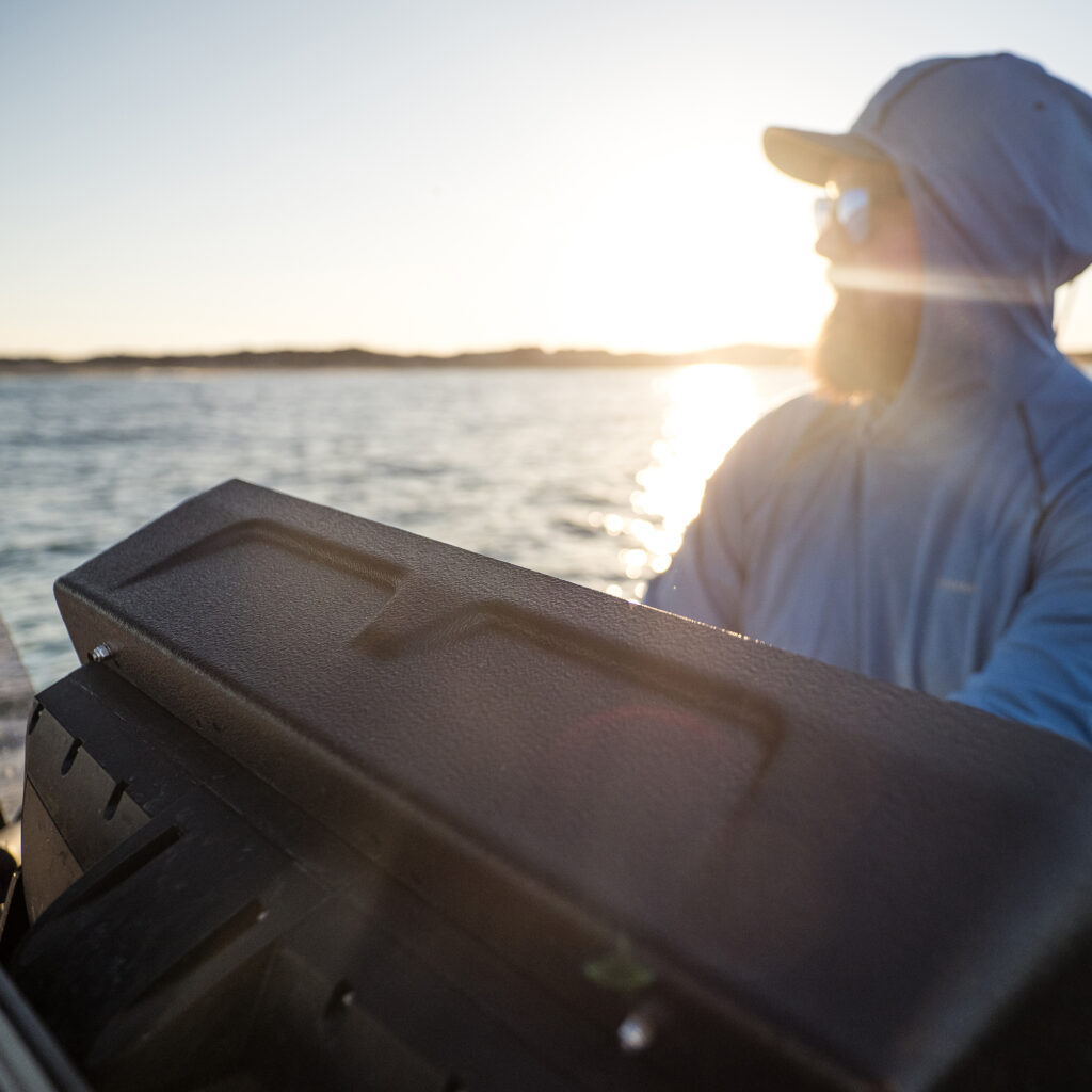 Rockin' a sleek hooded jacket, choice sunglasses, and the sharp B&G Vulcan visor, this legend takes charge of the boat with unshakeable confidence. The sun is blazing down as they glide smoothly over sparkling waters with distant lands just a blur in the background.