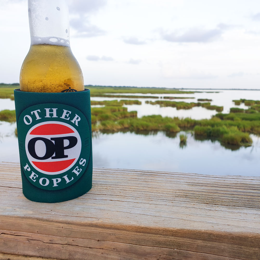 Nestled on the wooden railing, a bottle of beer chills out in its green OP Stubby Holder, soaking in the scenic vibe of the marshland.