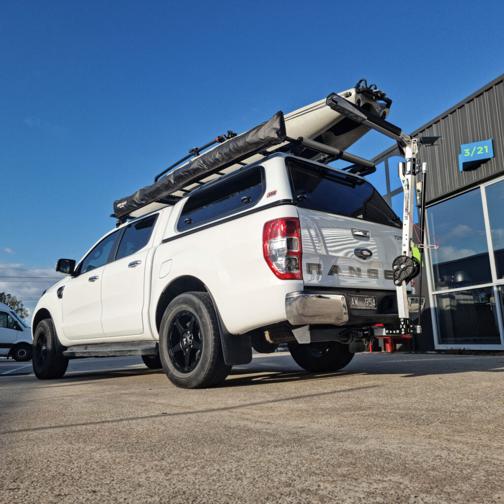 Parked on a smooth concrete surface close to a building, this white pickup truck is geared up and ready for adventure with its trusty Will-I-Loader. Perfectly designed for hauling kayaks and all your essential outdoor gear, this setup screams readiness and rugged exploration!.